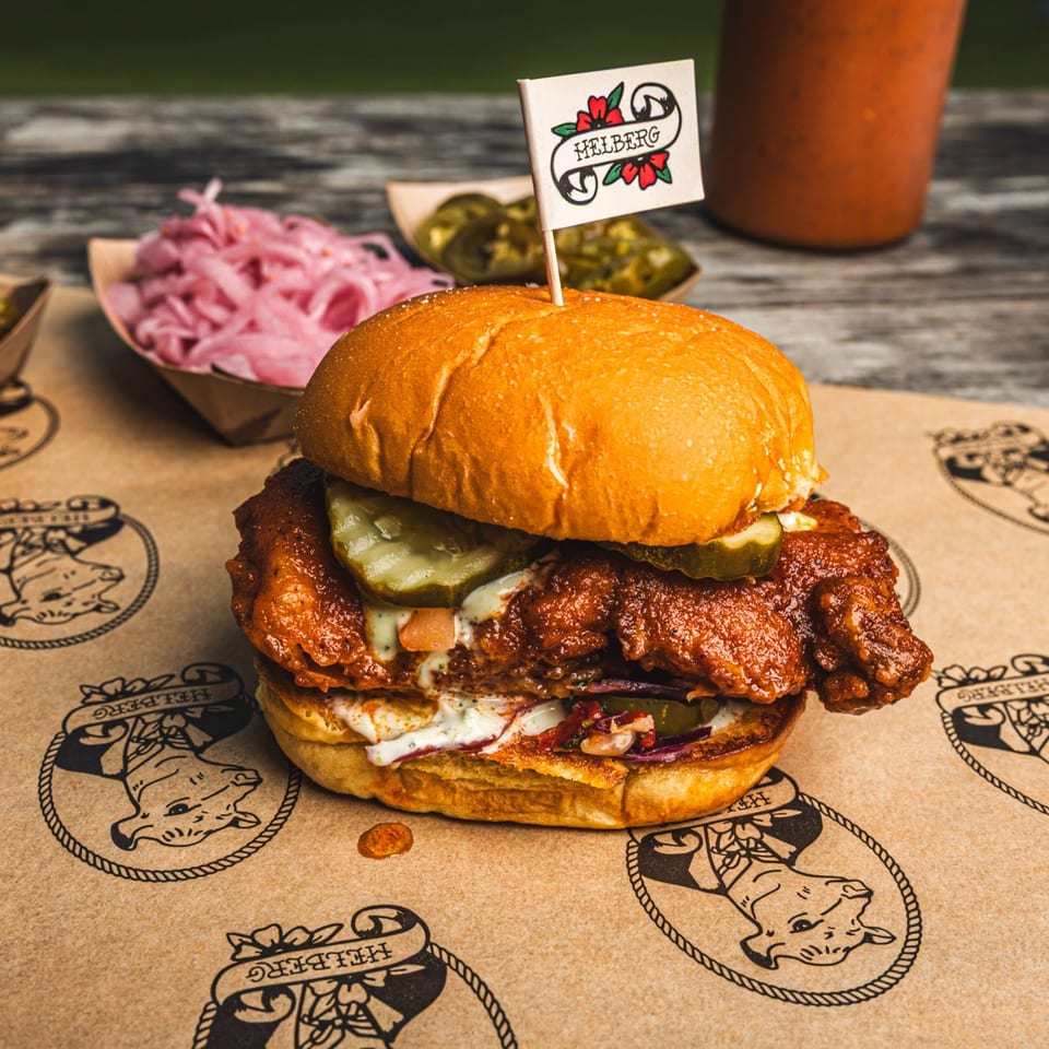 Crispy fried chicken sandwich with pickles and sauce on branded paper, accompanied by pickles, onions, jalapeños, and drinks.