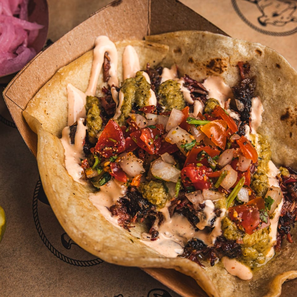 Loaded folded flatbread topped with grilled meat, creamy sauce, green salsa, diced tomatoes, onions, pickles, and pink pickled onions.