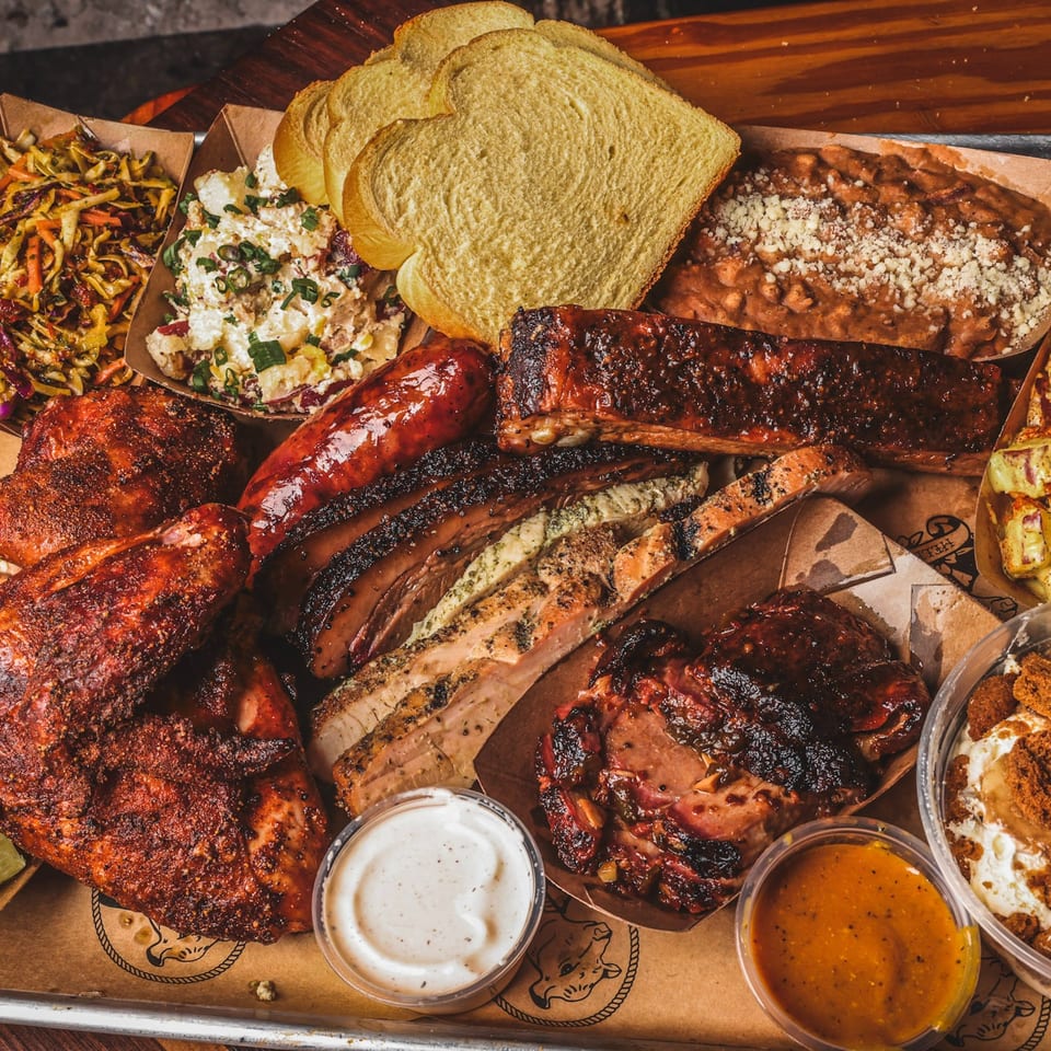 Overhead spread of barbecue meats, ribs, sausage, cornbread, beans, slaw, salads, sauces, and creamy side dishes on a wooden table.